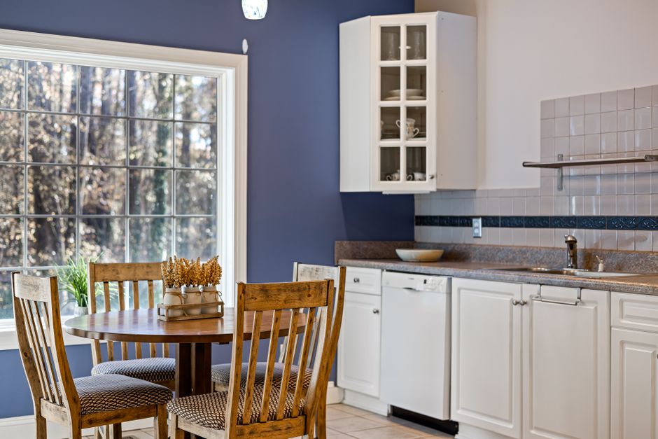 A bright kitchen with a round wooden dining table, white cabinets, and large window.